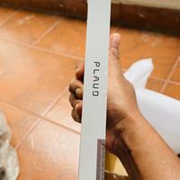 A person's hands holding a white rectangular box or package with "PLAUD" text printed on it, photographed indoors on a tiled floor