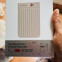 A person holding a white product box for the PLAUD NOTE, which is labeled as "The World's No.1 AI Voice Recorder" and shows an image of a slim, rectangular recording device with vertical lines on its surface.